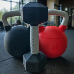 May include: A black 45-pound dumbbell with a white grip is centered between a black and a red kettlebell, each marked with the number 30. The weights are on a black gym floor.