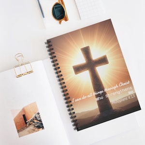 Puede incluir: Cuaderno de espiral con una cruz sobre un fondo de rayos de sol. La portada incluye el texto "I can do all things through Christ Who Strengthens Me" y "Philippians 4:13". El cuaderno está abierto sobre una superficie blanca.