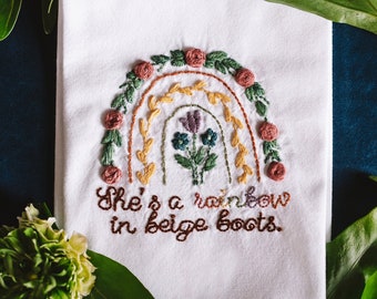 May include: White tea towel with an embroidered rainbow and the text "She's a rainbow in beige boots."