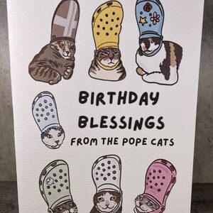 Puede incluir: Una tarjeta de felicitaci&oacute;n con una ilustraci&oacute;n humor&iacute;stica de gatos que llevan diferentes estilos de zapatos Crocs como sombreros. El texto de la tarjeta dice "Birthday Blessings from the Pope Cats".