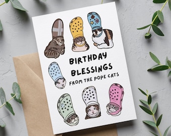 May include: A greeting card with a humorous illustration of cats wearing colorful Crocs as hats. The text reads "Birthday Blessings from the Pope Cats".