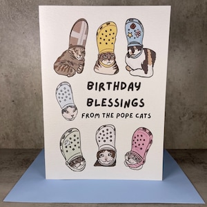 Puede incluir: Una tarjeta blanca con un saludo de cumplea&ntilde;os que dice "Birthday Blessings from the Pope Cats". La tarjeta presenta seis ilustraciones de gatos con Crocs de diferentes colores.