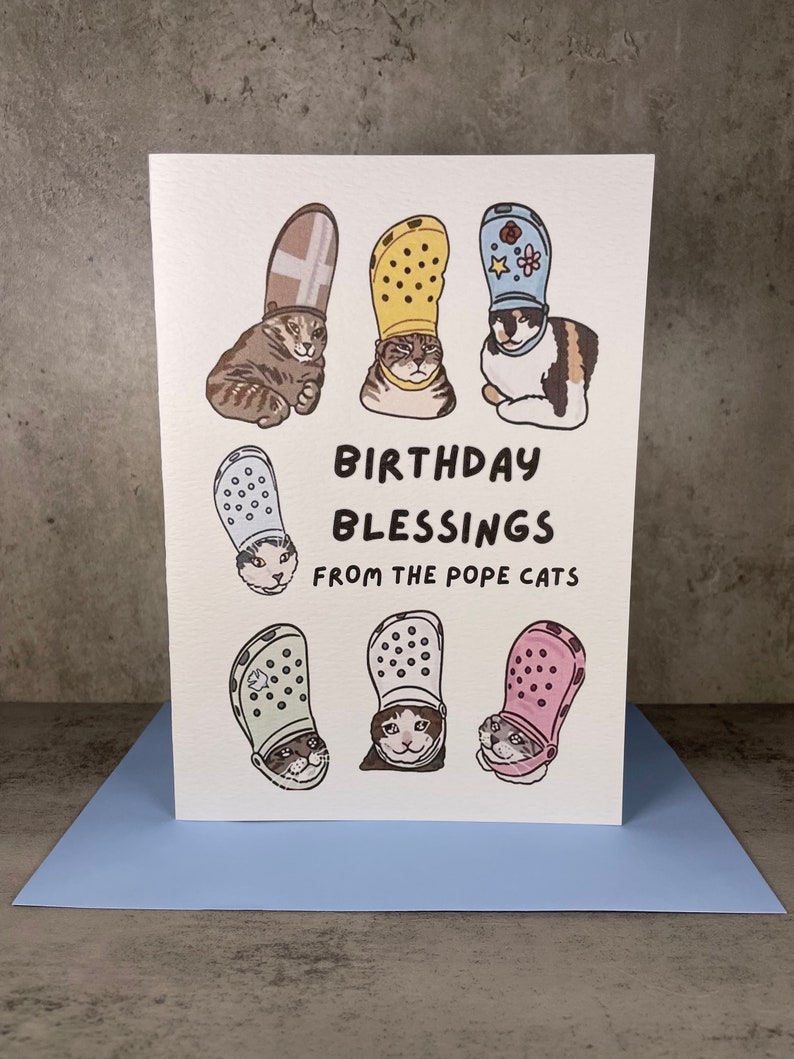Puede incluir: Una tarjeta blanca con un saludo de cumplea&ntilde;os que dice "Birthday Blessings from the Pope Cats". La tarjeta presenta seis ilustraciones de gatos con Crocs de diferentes colores.
