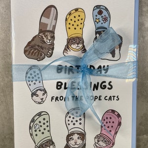 Puede incluir: Una tarjeta de cumplea&ntilde;os con una cinta azul atada alrededor. La tarjeta presenta una ilustraci&oacute;n de dibujos animados de gatos con Crocs coloridos. El texto de la tarjeta dice "Birthday Blessings from the Croc Cats".