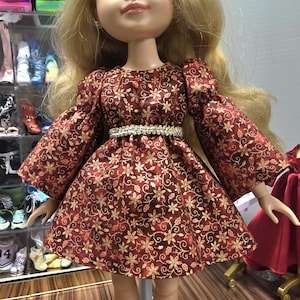 May include: A doll with long blonde hair wearing a burgundy dress with gold floral patterns. The dress has long sleeves and a jeweled belt. The doll is standing on a white stand, with a background of shelves filled with shoes.