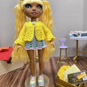 Könnte beinhalten: Eine Puppe mit langen blonden Haaren, einem blau-weißen Häkelkleid und bunten Plateau-Sneakers. Sie trägt eine gelbe Häkeljacke mit einem blauen Knopf. Die Puppe steht auf einer Plattform mit dem Wort "Rainbow" darauf. Die Puppe hat braune Haut und grüne Augen. Sie trägt ein gelbes Häkelhaarband mit einem Sternmuster. Die Puppe hat die Zahl "19" auf ihrer Wange.