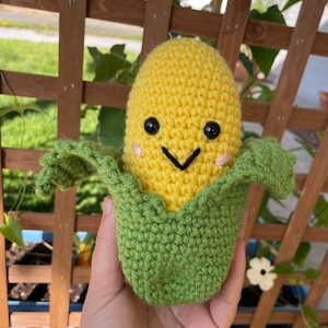 May include: A crocheted corn on the cob plush toy with a smiling face, yellow kernels, and green husks.