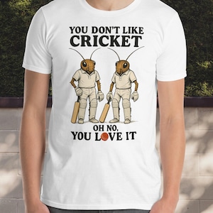 以下が含まれることがあります： 「YOU DON'T LIKE CRICKET」の文字と、クリケットをする2匹の昆虫のイラストがプリントされた白いTシャツ。「OH NO, YOU LOVE IT」の文字がイラストの下にあります。昆虫はクリケットのユニフォームを着て、バットを持っています。