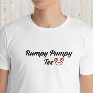 Puede incluir: Camiseta blanca con texto negro que dice "Rumpy Pumpy Tee" y una cara sonriente roja.