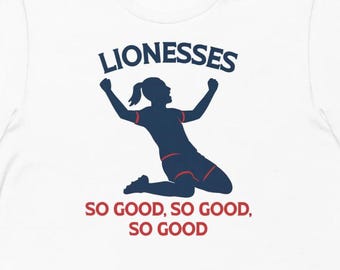 May include: White t-shirt with a graphic of a silhouette of a woman celebrating, with the word "LIONESSES" above and the phrase "SO GOOD, SO GOOD, SO GOOD" below. The graphic is in navy blue and red.