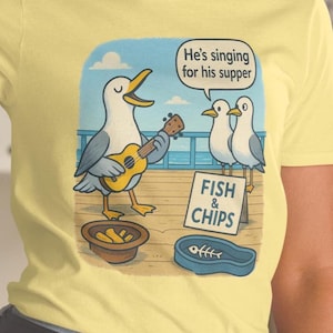 Può includere: T-shirt gialla con un disegno di un gabbiano che suona l'ukulele. Il design include il testo "He's singing for his supper" e un cartello con la scritta "Fish & Chips". Sono presenti anche altri due gabbiani.
