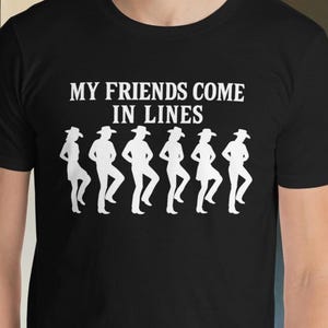 May include: Black t-shirt with the text "MY FRIENDS COME IN LINES" above a white graphic of line dancers. The dancers are wearing hats and are in a row. The shirt is short-sleeved.