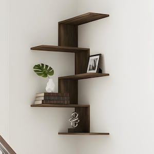 May include: A dark brown, zig-zag corner shelf unit on a white wall, holding decorative items. These include a white vase with a green leaf, books, a framed photo, and a sculpture. The staircase has dark wood steps and a cream carpet runner.