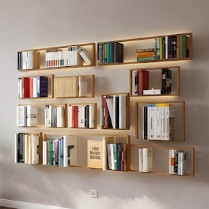 May include: A modern living room with a wall-mounted bookshelf system. The shelves are made of light wood and are filled with books of various sizes and colors. The room also features a teal armchair, a potted plant, and a fluffy white rug.