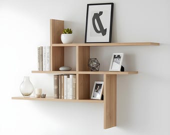 May include: A modern, light-colored wooden wall shelf unit with multiple compartments and shelves. The shelves display books, decorative objects, and framed photos. A gray armchair and side table are in the foreground, creating a living room setting.