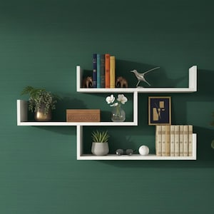 以下が含まれることがあります： 濃い緑色の壁に飾られた、装飾品付きの白いフローティングシェルフ。本、植物、小さな彫刻、額縁、木箱が置かれています。シェルフはユニークな幾何学模様に配置されています。