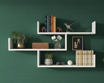 May include: White floating shelves with decorative items against a dark green wall. The shelves hold books, plants, small sculptures, a framed picture, and a wooden box. The shelves are arranged in a unique, geometric design.