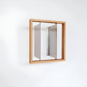 May include: A modern wall art piece featuring a light brown wooden frame with three vertical, rectangular, silver-colored panels. The panels are arranged within the frame, creating a minimalist, geometric design. The artwork is mounted on a white wall.
