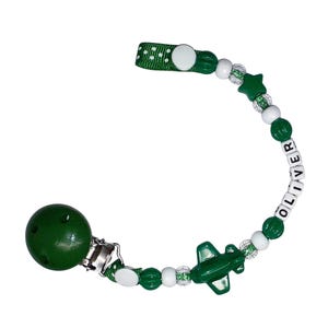 May include: A green and white pacifier clip with the name "OLIVER" spelled out in white letter beads. The clip features a green airplane charm, a green star, and a green and white polka dot ribbon.
