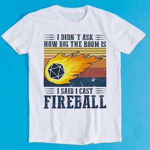 Könnte beinhalten: Ein weißes Kurzarm-T-Shirt mit einer Grafik eines zwanzigseitigen Würfels in einem gelben und orangen Feuerball. Das Design enthält horizontale Streifen in Sonnenuntergangsfarben und den Text: I DIDN'T ASK HOW BIG THE ROOM IS I SAID I CAST FIREBALL.