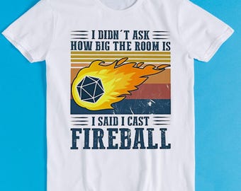 Könnte beinhalten: Ein weißes Kurzarm-T-Shirt mit einer Grafik eines zwanzigseitigen Würfels in einem gelben und orangen Feuerball. Das Design enthält horizontale Streifen in Sonnenuntergangsfarben und den Text: I DIDN'T ASK HOW BIG THE ROOM IS I SAID I CAST FIREBALL.