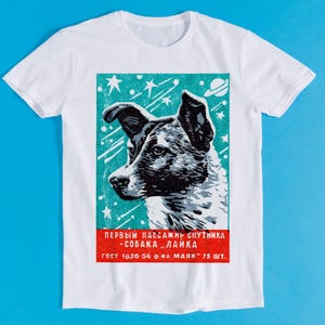 以下が含まれることがあります： 宇宙にいる犬のグラフィックが特徴の白いTシャツ。犬は白黒で、星と惑星のあるターコイズブルーの背景に配置されています。下部に赤と白のテキストがあります。