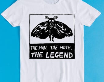 May include: A white short-sleeved t-shirt featuring a black and white graphic of a moth centered in a square frame. Below the moth, a black rectangular block contains the white text: THE MAN, THE MOTH, THE LEGEND, printed in a bold, hand-drawn style.