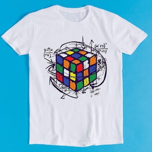 Może przedstawiać: Biała koszulka z kolorowym nadrukiem kostki Rubika i otaczającymi ją równaniami i symbolami matematycznymi.