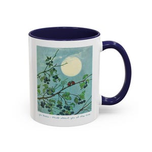 Könnte beinhalten: Weißer Keramikbecher mit marineblauem Innenraum und Henkel. Der Becher zeigt eine skurrile Illustration eines Vollmonds, eines Astes mit Beeren und zwei Marienkäfern. Der Text "you know; I think about you all the time" ist auf dem Becher aufgedruckt.