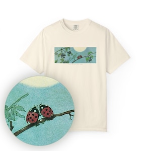 以下が含まれることがあります： クリーム色のTシャツ。自然をテーマにしたグラフィックが特徴です。グラフィックは、ベリーのある枝、太陽、2匹のテントウムシを描いています。「you can't kill my vibe」というテキストが画像の下に印刷されています。