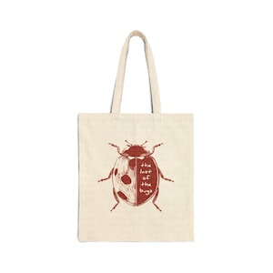 May include: A beige canvas tote bag featuring a dark red graphic of a ladybird. The left side of the ladybird shows spots, while the right side contains the text "the last of the bugs" written in a simple, hand-drawn style font across the wing.
