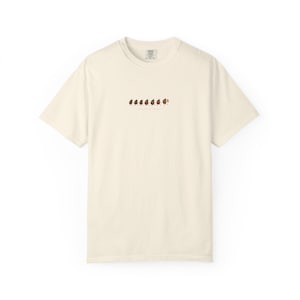 以下が含まれることがあります： クルーネックと半袖のクリーム色のTシャツ。シャツには、いくつかの小さな葉が付いた茶色の形がいくつか水平にデザインされています。デザインの下には「in the air」という文字があります。シャツは柔らかい素材で作られています。