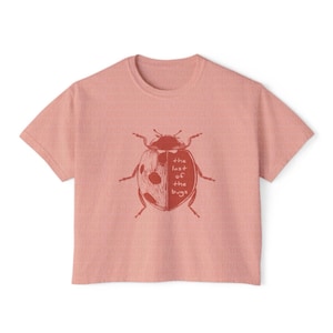 May include: A dusty rose short-sleeved t-shirt featuring a dark red graphic of a ladybug on the chest. The ladybug illustration includes the text "the last of the bugs" written in a simple, handwritten font across the center of its shell.