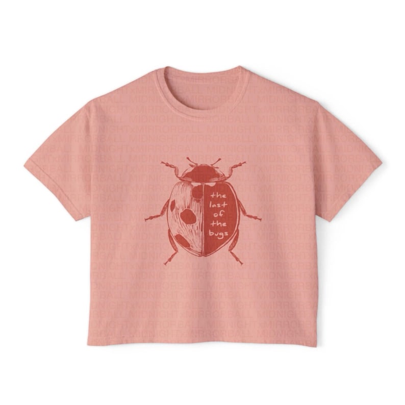 May include: A dusty rose short-sleeved t-shirt featuring a dark red graphic of a ladybug on the chest. The ladybug illustration includes the text "the last of the bugs" written in a simple, handwritten font across the center of its shell.