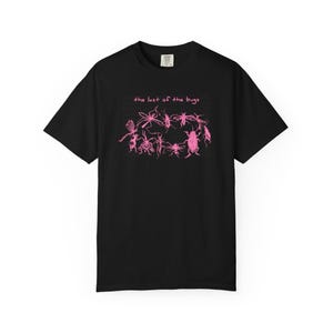 May include: A black short-sleeved t-shirt featuring a pink graphic of various insects arranged in a circle. Above the insects, the text reads "the last of the bugs" in a pink handwritten font. The shirt has a crew neckline and a standard fit.