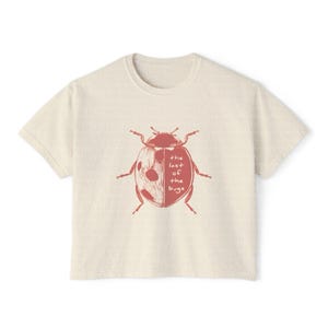 May include: A cream-colored short-sleeve t-shirt featuring a red graphic of a ladybug on the chest. The ladybug illustration includes the text 'the last of the bugs' written in a simple, handwritten font across the right side of its shell.
