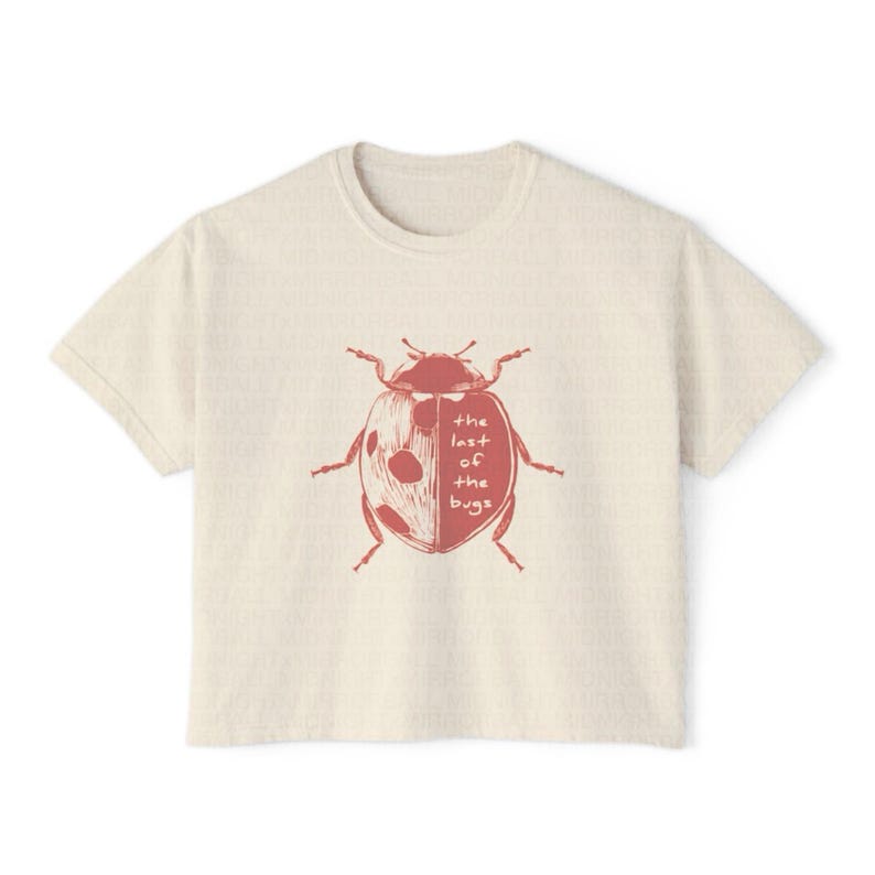 May include: A cream-colored short-sleeve t-shirt featuring a red graphic of a ladybug on the chest. The ladybug illustration includes the text 'the last of the bugs' written in a simple, handwritten font across the right side of its shell.
