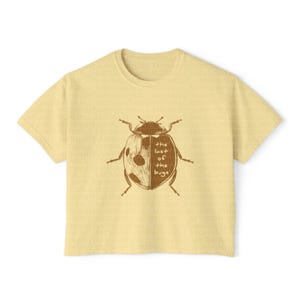 May include: A pale yellow short-sleeve t-shirt featuring a brown graphic of a ladybug on the front. The ladybug illustration is split down the middle, with the text "the last of the bugs" printed in a simple font over the right side of the insect.