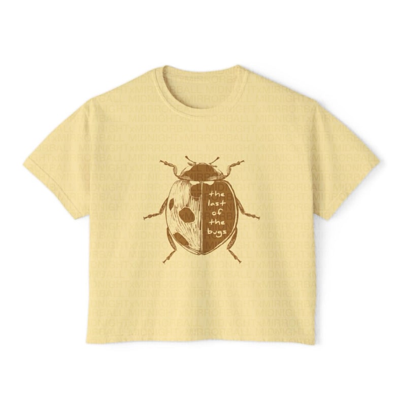 May include: A pale yellow short-sleeve t-shirt featuring a brown graphic of a ladybug on the front. The ladybug illustration is split down the middle, with the text "the last of the bugs" printed in a simple font over the right side of the insect.