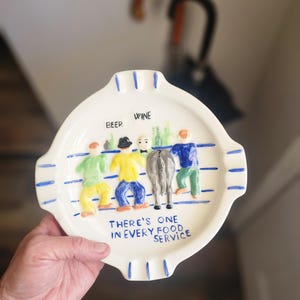 May include: Cream-colored ceramic ashtray with a humorous illustration. The design features figures labeled "Beer" and "Wine" with a donkey. The text reads "There's one in every food service." Blue stripes and accents.