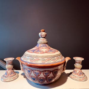 May include: A ceramic serving dish with a lid and two matching candle holders. The dish and candle holders are decorated with geometric patterns in shades of blue, pink, and orange. The dish has handles and a decorative knob on the lid.