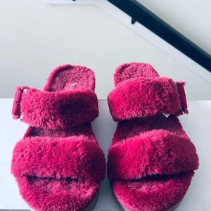 May include: A pair of fuchsia pink faux fur slides featuring two wide straps across the top. Each sandal includes a matching fuchsia pink adjustable buckle on the outer side of the top strap. The footbed and straps are covered in plush, textured material.
