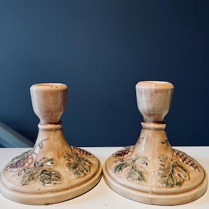 May include: Two ceramic candle holders with a beige glaze. Each holder features a base decorated with grape leaves and clusters. The leaves are green and the grapes are purple. The candle holders are on a white surface with a dark blue background.