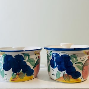 May include: Two ceramic bowls with lids, featuring hand-painted designs of blue grapes, green leaves, and red and yellow fruit. The bowls have a white base with a blue rim and a white handle on the lid. These decorative bowls are ideal for kitchen decor.