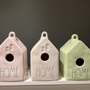May include: Three ceramic birdhouses in pastel colors: pink, off-white, and green. Each house-shaped item has a circular opening and a small loop at the top. The word "HOME" is written in black script on the front of each birdhouse.
