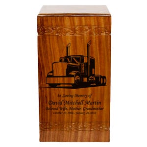 May include: A wooden urn with a truck design. The urn is rectangular with a light brown finish and a carved floral design at the top. The front features a black illustration of a semi-truck and the text "In Loving Memory of David Mitchell Martin".