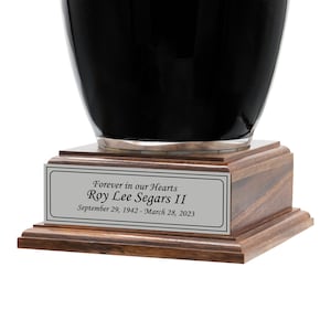 May include: A black urn with a silver rim sits atop a wooden base. A rectangular silver plaque on the base reads "Forever in our Hearts" and the name "Roy Lee Segars II" with the dates September 29, 1942 - March 28, 2023.