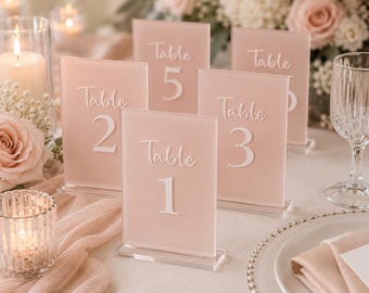 May include: Pink acrylic table number signs with white lettering, numbered 1 through 5. The signs are displayed on a table with a candle, flowers, and a wine glass. The signs are for a wedding or event.