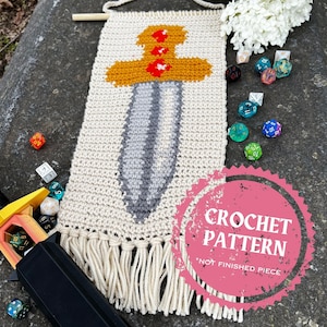 May include: Crochet pattern for a sword wall hanging. The sword is gray and yellow with red accents. The pattern is not a finished piece. The image also includes dice and a wooden stick.
