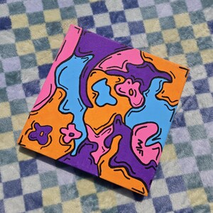 Peut inclure: Une peinture abstraite colorée sur toile. L'œuvre présente un mélange vibrant de formes orange, rose, bleu et violet, soulignées en noir. Les initiales "MF" sont visibles en bas à droite. La toile est présentée sur un fond à carreaux.